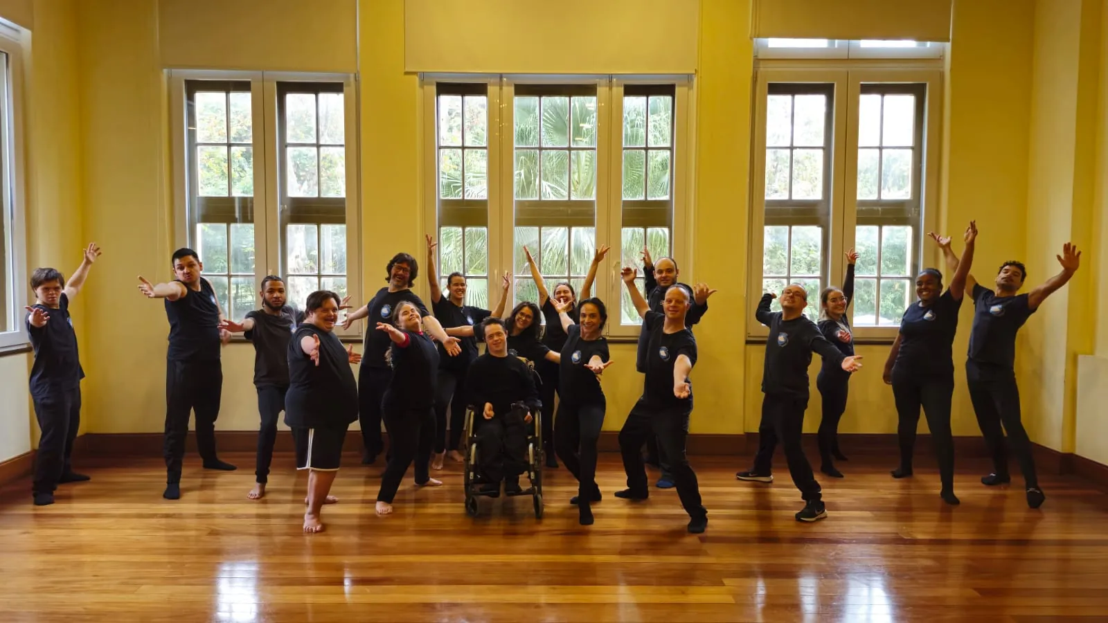 Turma da oficina de dança de braços erguidos no salão, sorrindo para a câmera.