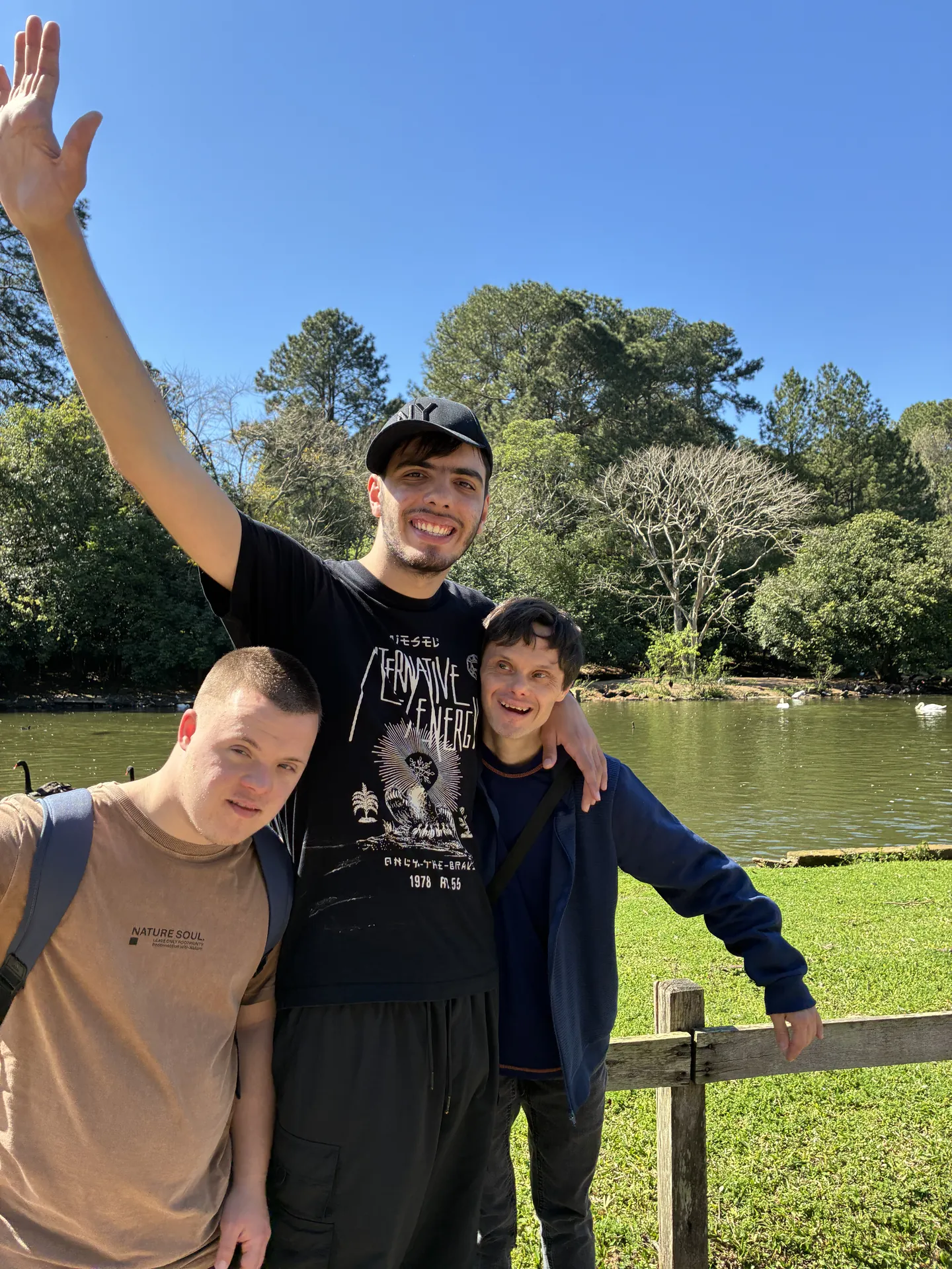 Trio de participantes abraçados num parque, com o lago ao fundo.