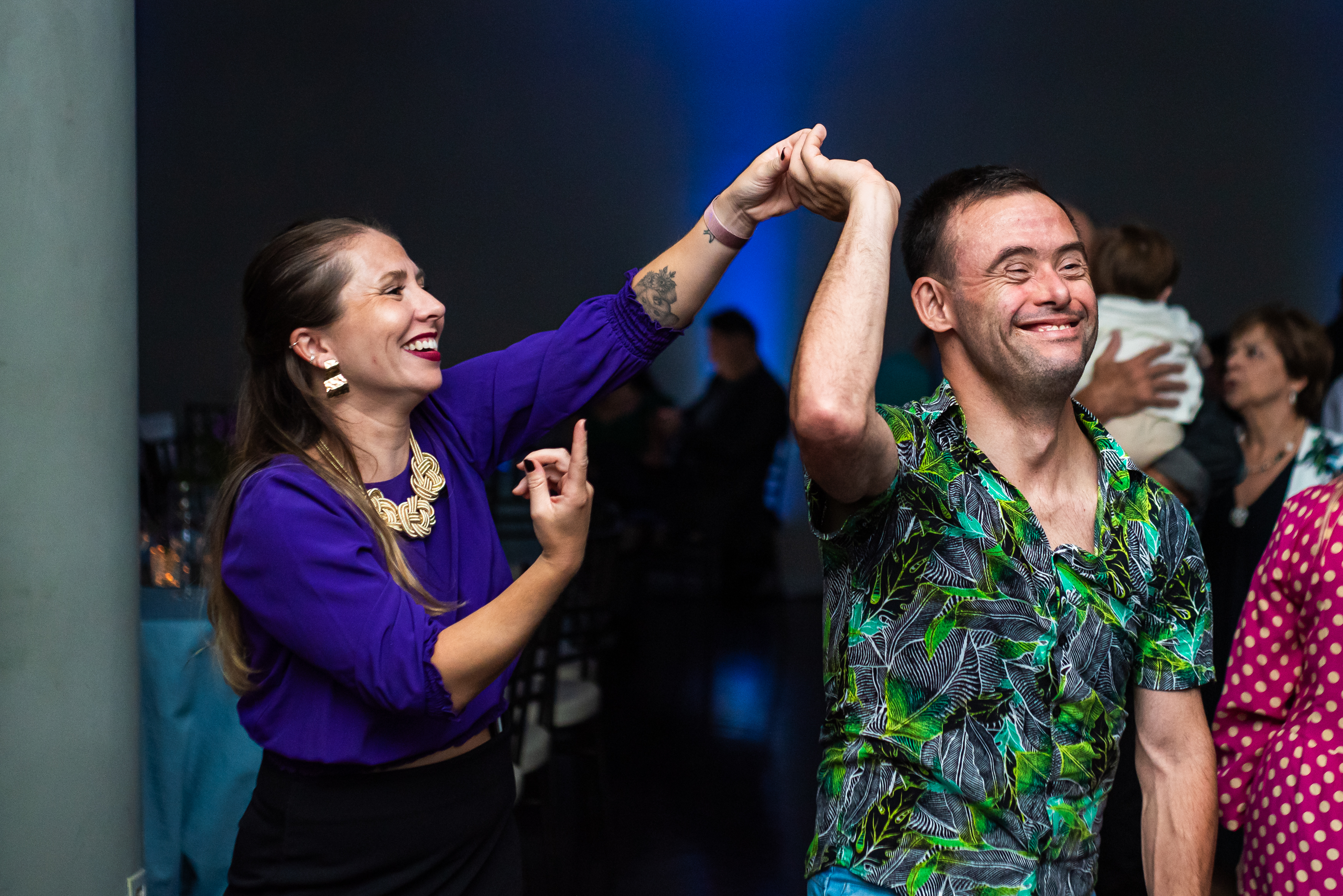 Homem com síndrome de Down de camisa estampada dança de mãos dadas com uma mulher, os dois sorrindo, em uma festa.