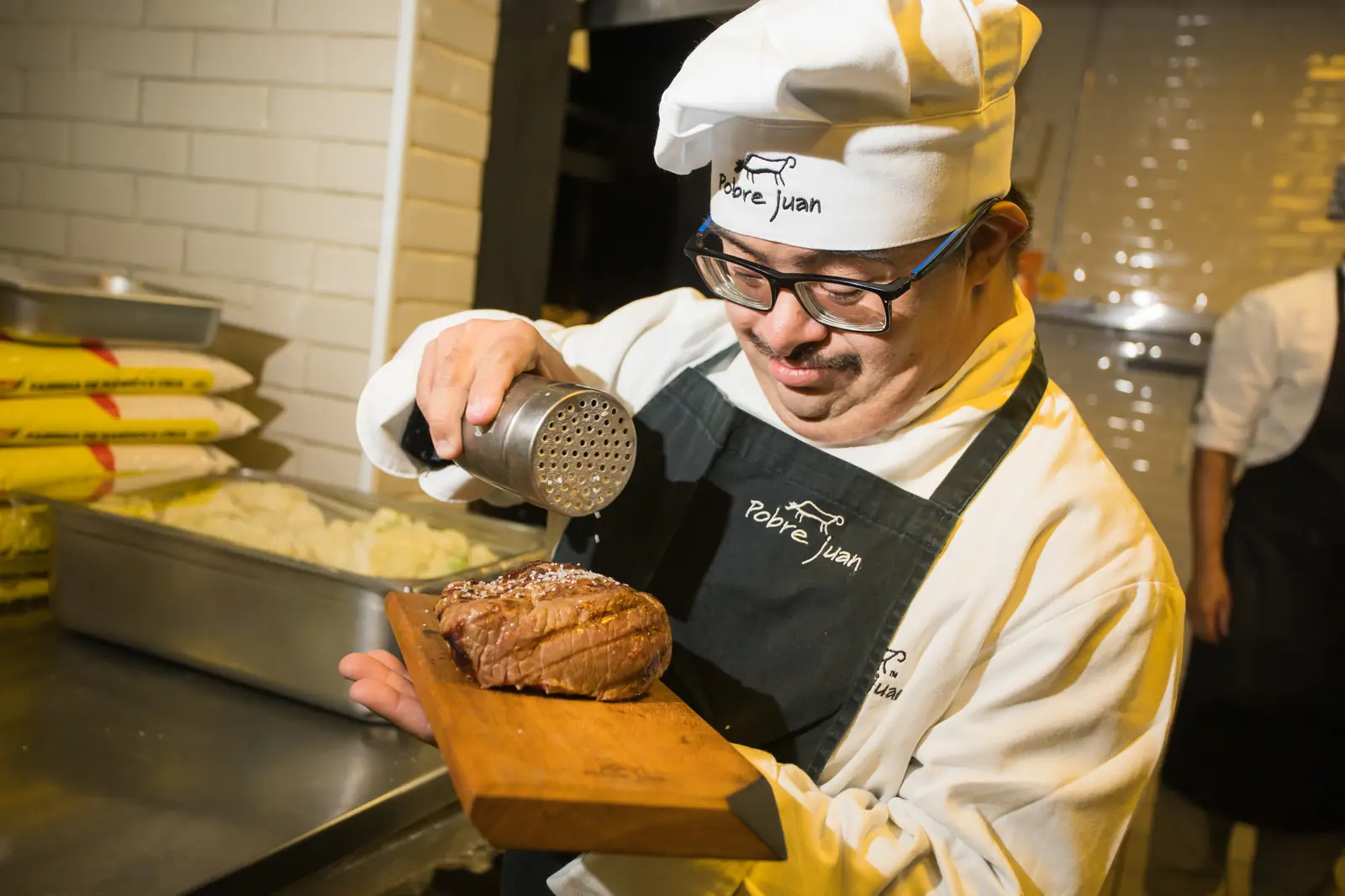 Cozinheiro de touca e avental salga um corte de carne sobre uma tábua de madeira, na cozinha de um restaurante.