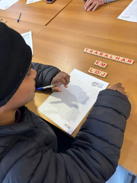Participante escreve em uma folha pautada; sobre a mesa, peças de letras formam a frase trabalhar em loja.