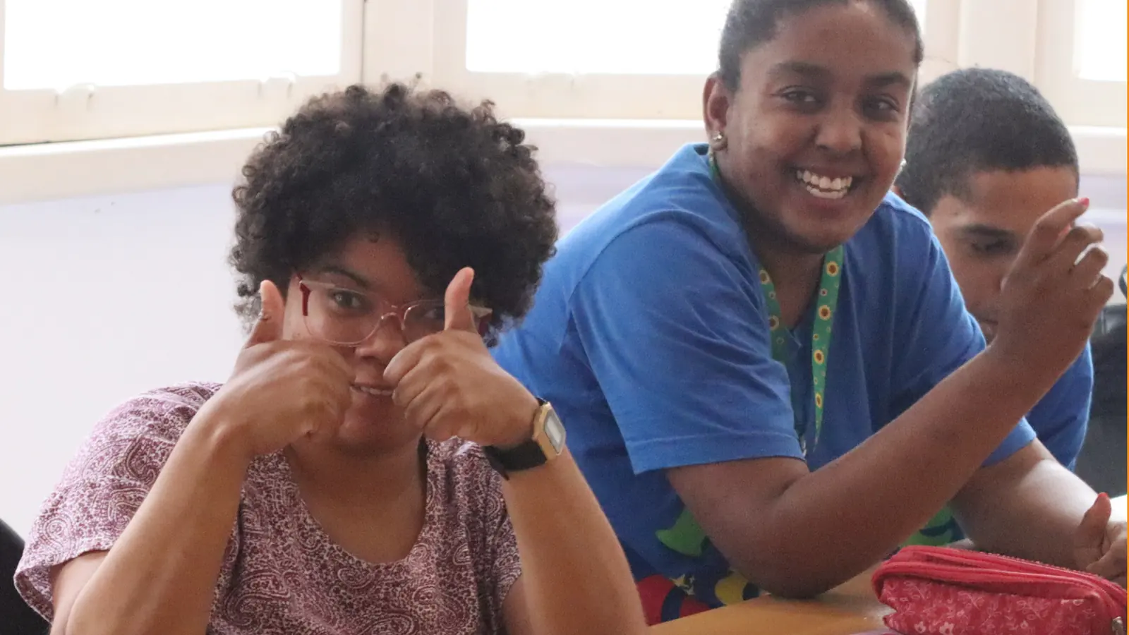 Duas participantes sorriem para a câmera em sala de aula; uma delas faz sinal de positivo com as duas mãos.