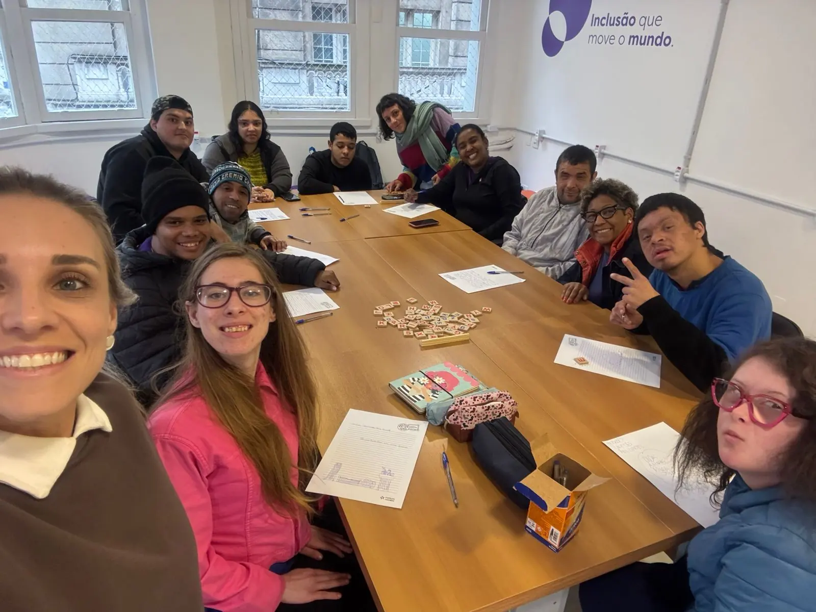 Turma reunida em volta de uma mesa com folhas de atividade e peças de letras, sorrindo para a foto.