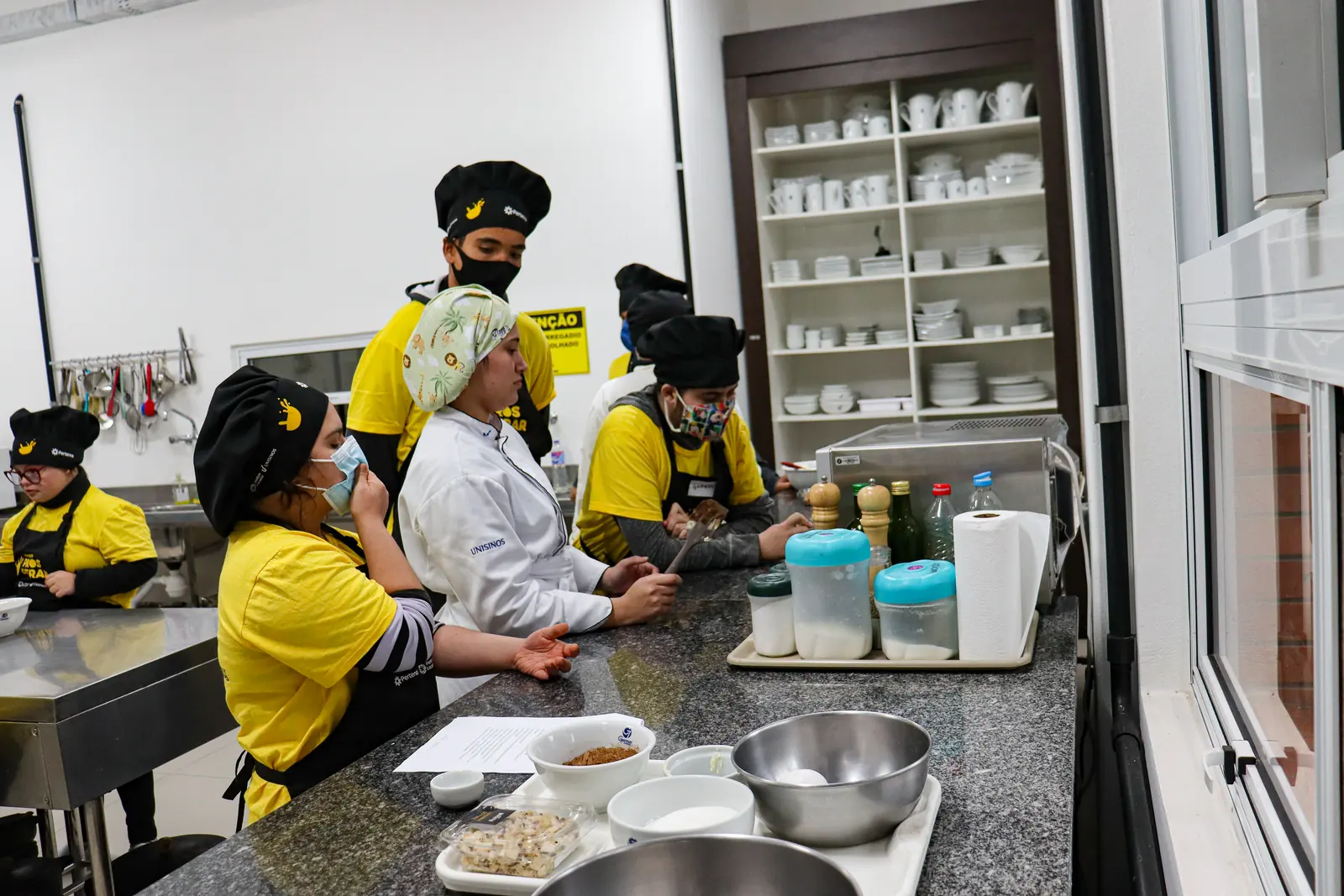 Aula prática na cozinha, com participantes e instrutores em volta da bancada.