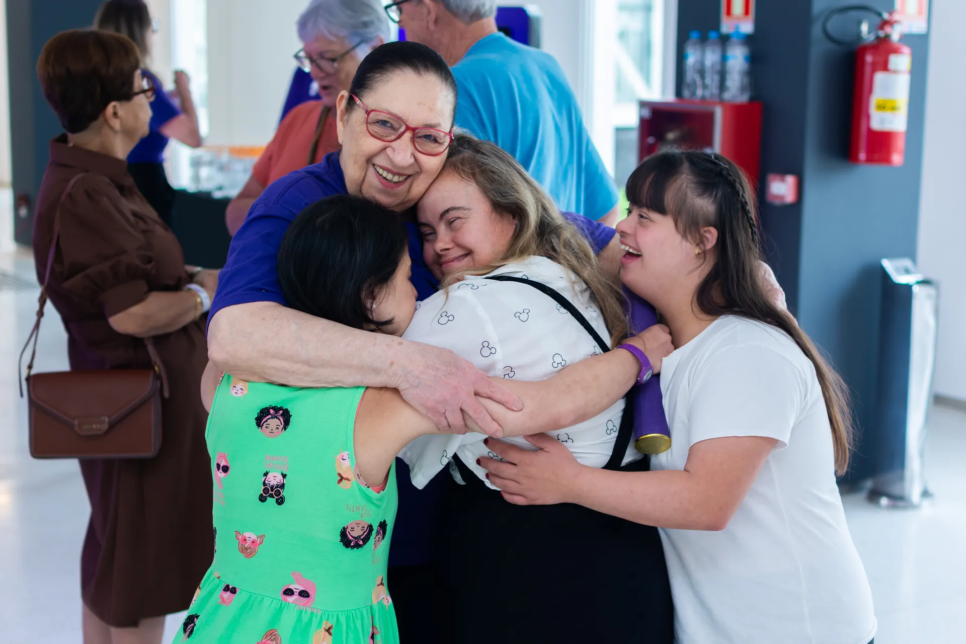 Quatro mulheres, entre elas duas jovens com síndrome de Down, se abraçam sorrindo num encontro do Pertence.